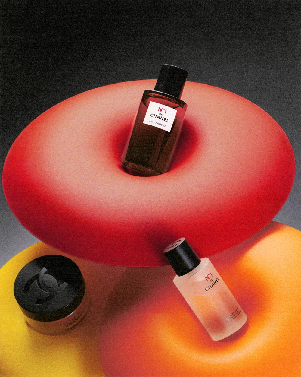 Product photography of three Chanel fragrances arranged on a large red spherical form with yellow and orange geometric shapes in the background.