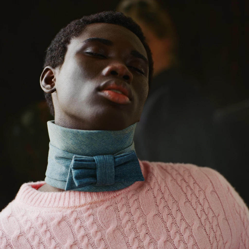 Close-up portrait photograph of a person wearing a pink cable-knit sweater and a teal blue bow-tie neck accessory, shot against a dark blurred background.