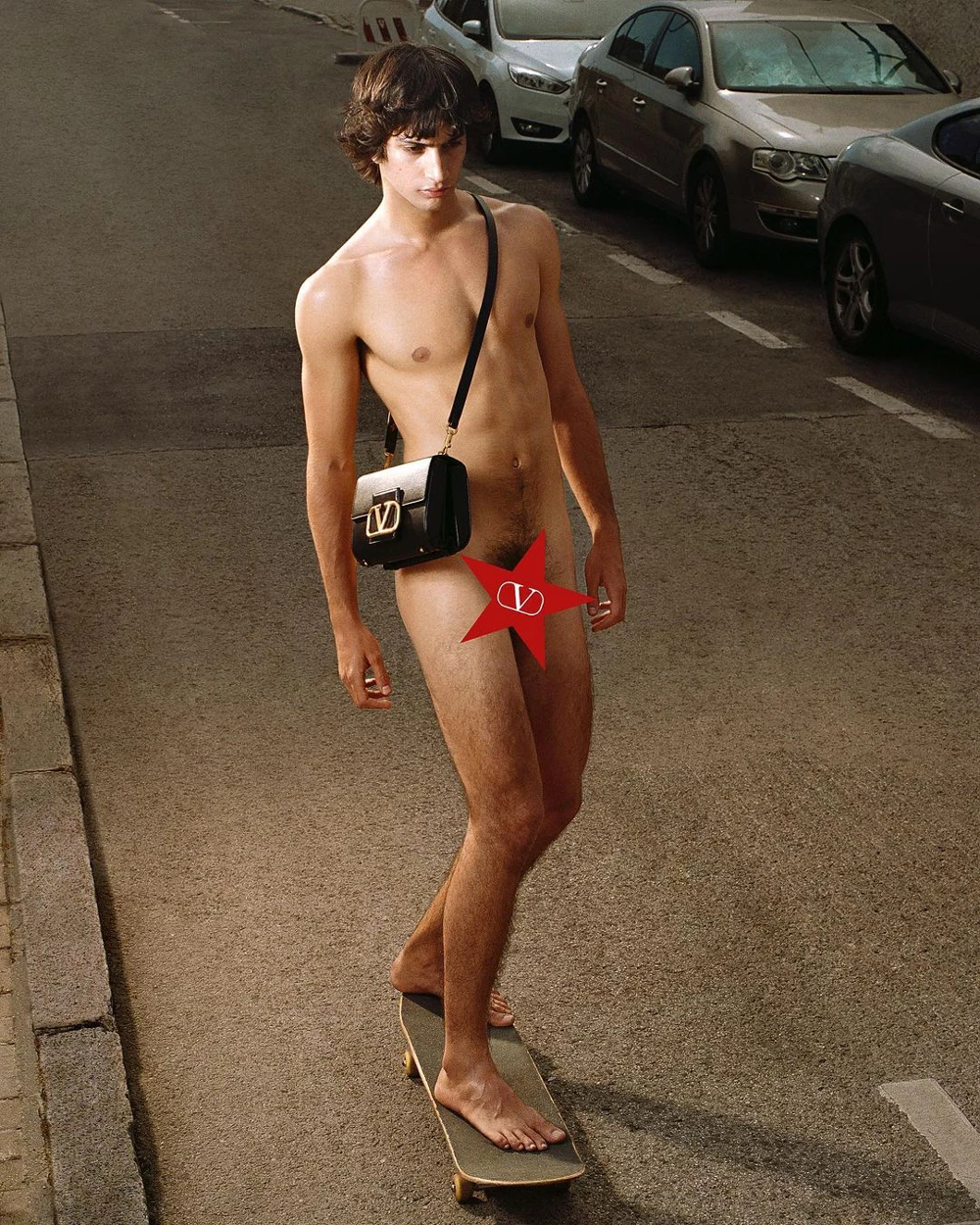 Color photograph of a shirtless male figure on a skateboard in a parking lot, wearing a black and white designer shoulder bag and red star-shaped censoring element.