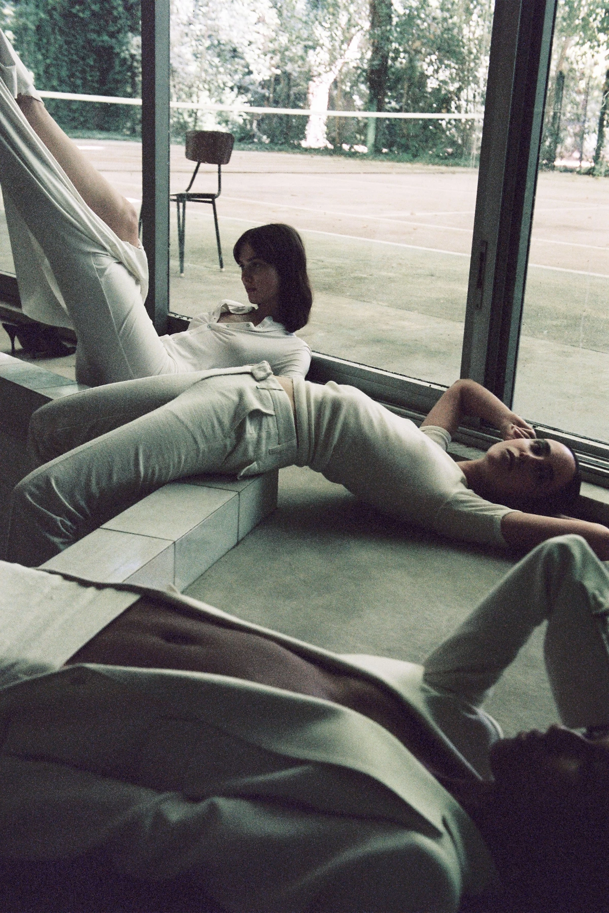Film photograph of two people relaxing on a concrete bench beside large windows overlooking a courtyard with trees and modernist furniture.